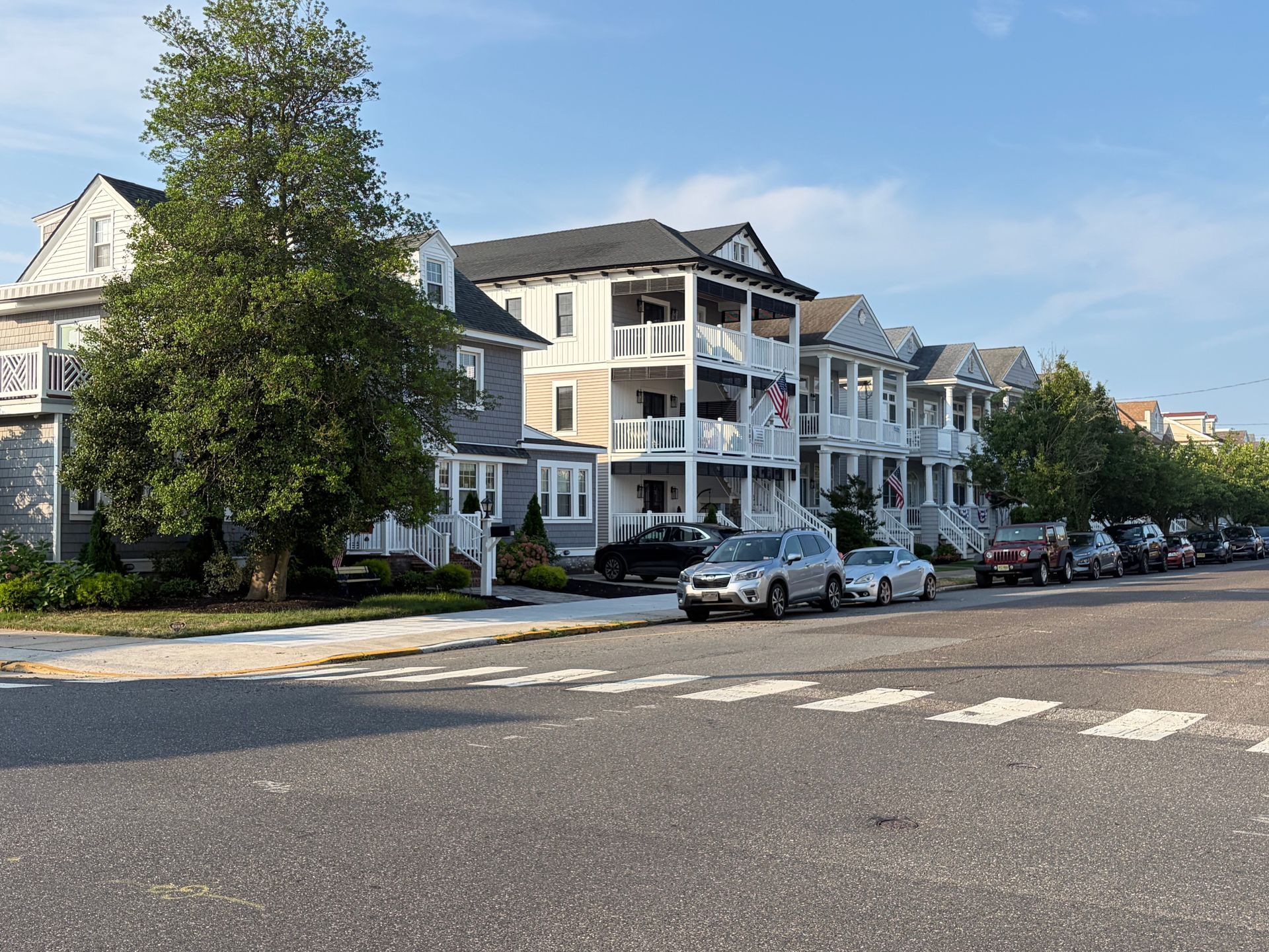 Row of white apartment buildings along a tree-lined street with parked cars and a crosswalk