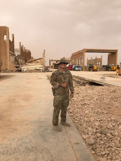 Cole Gasperini in uniform standing near partially destroyed buildings in a dusty, outdoor setting. Cole Gasperini in uniform standing near partially destroyed buildings in a dusty, outdoor setting.