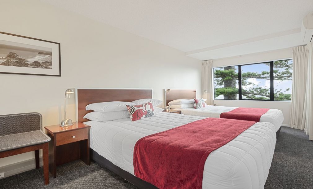 Bright hotel room with two neatly made beds, red throws, and a window view of trees and water