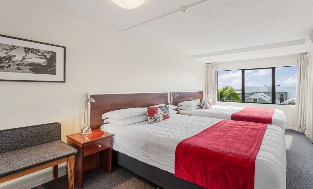 Bright hotel room with two beds, red runner, nightstand, and large window with mountain view