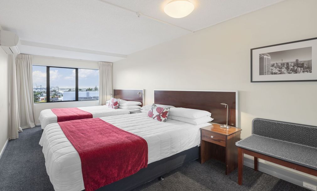 Bright hotel room with two twin beds, red bed runners, a window view, and a bench by the wall