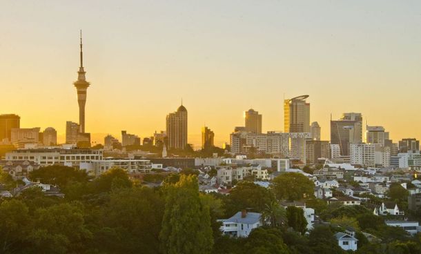 Sunset skyline with tall city buildings and a prominent tower above green trees