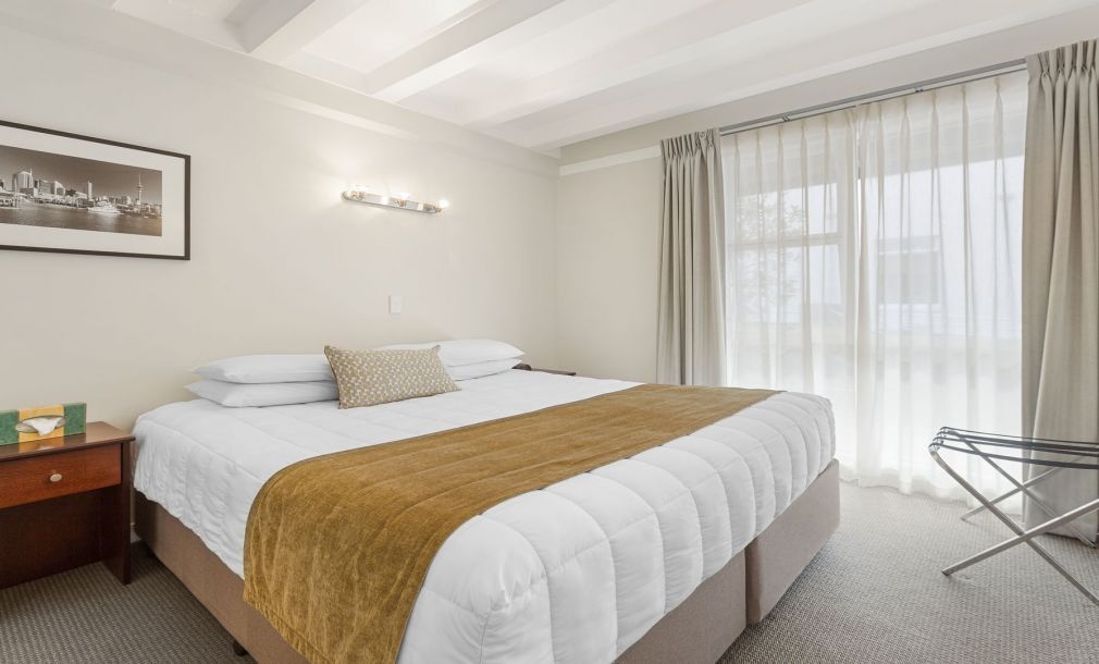 Bright bedroom with a large bed, mustard throw, bedside table, and sheer curtains by a window.