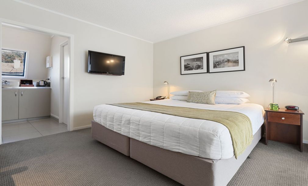 Modern bedroom with a large bed, wall-mounted TV, bedside tables, and an open doorway to a kitchenette.