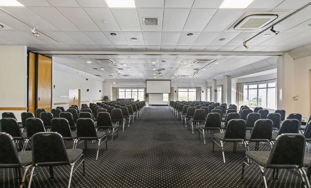 Empty conference room with rows of black chairs facing a projection screen and large windows.