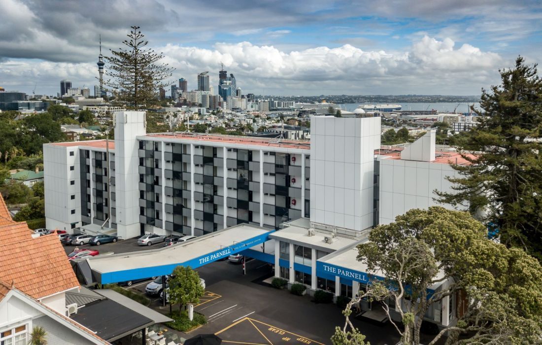 White hotel building with blue awning in front of a city skyline under cloudy skies