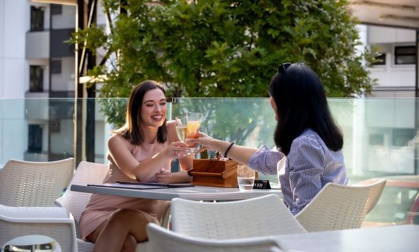 Two women clink drinks at an outdoor café table with laptops and coffee in a sunny patio setting.