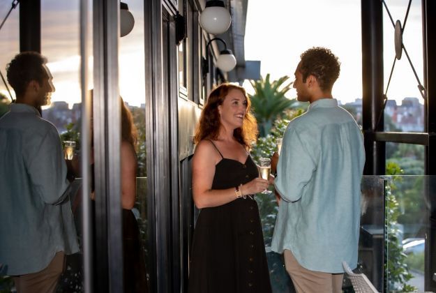 Two people talking on a balcony at sunset, one holding a drink, with city views behind them