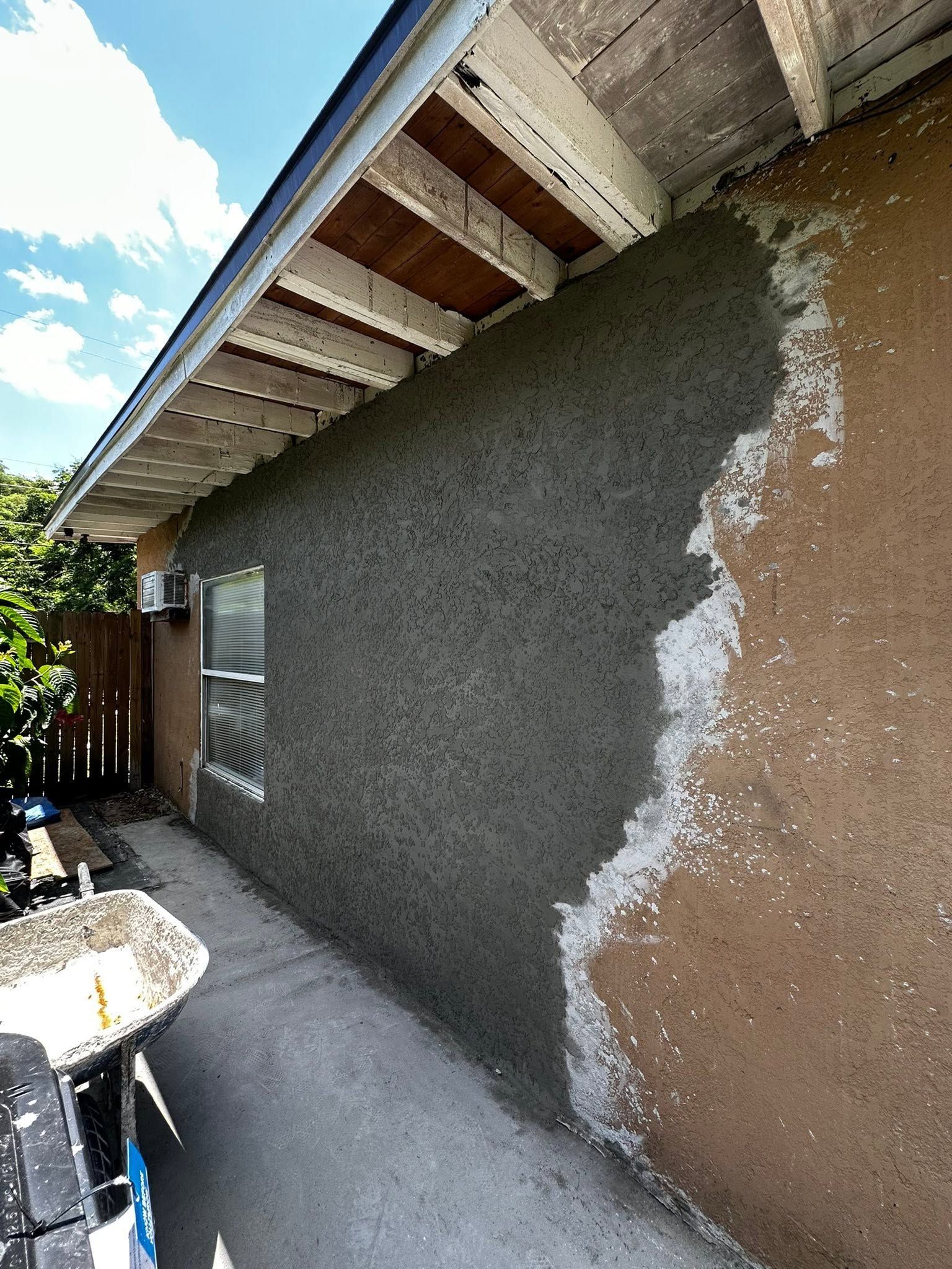Exterior wall under construction with gray stucco and exposed brown patch beneath a roof eave