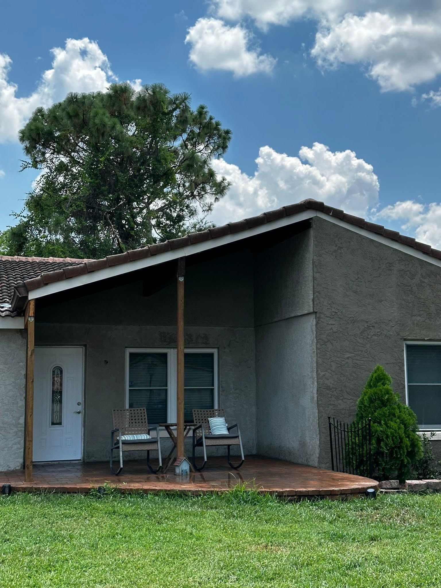 Small house with a porch, patio chairs, and a green lawn under a partly cloudy sky.