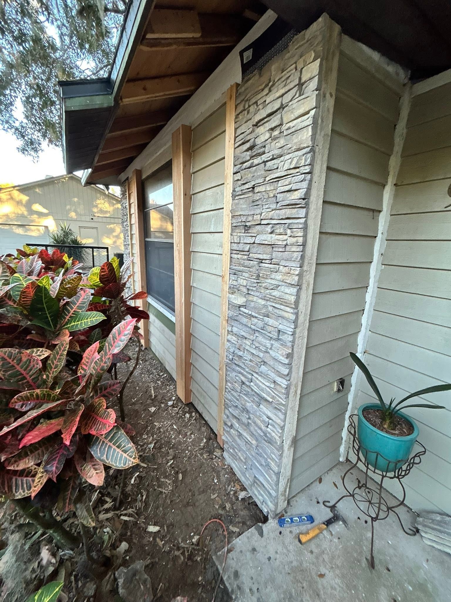 Narrow side yard beside a house with textured wall panels, plants, and a blue hanging pot.