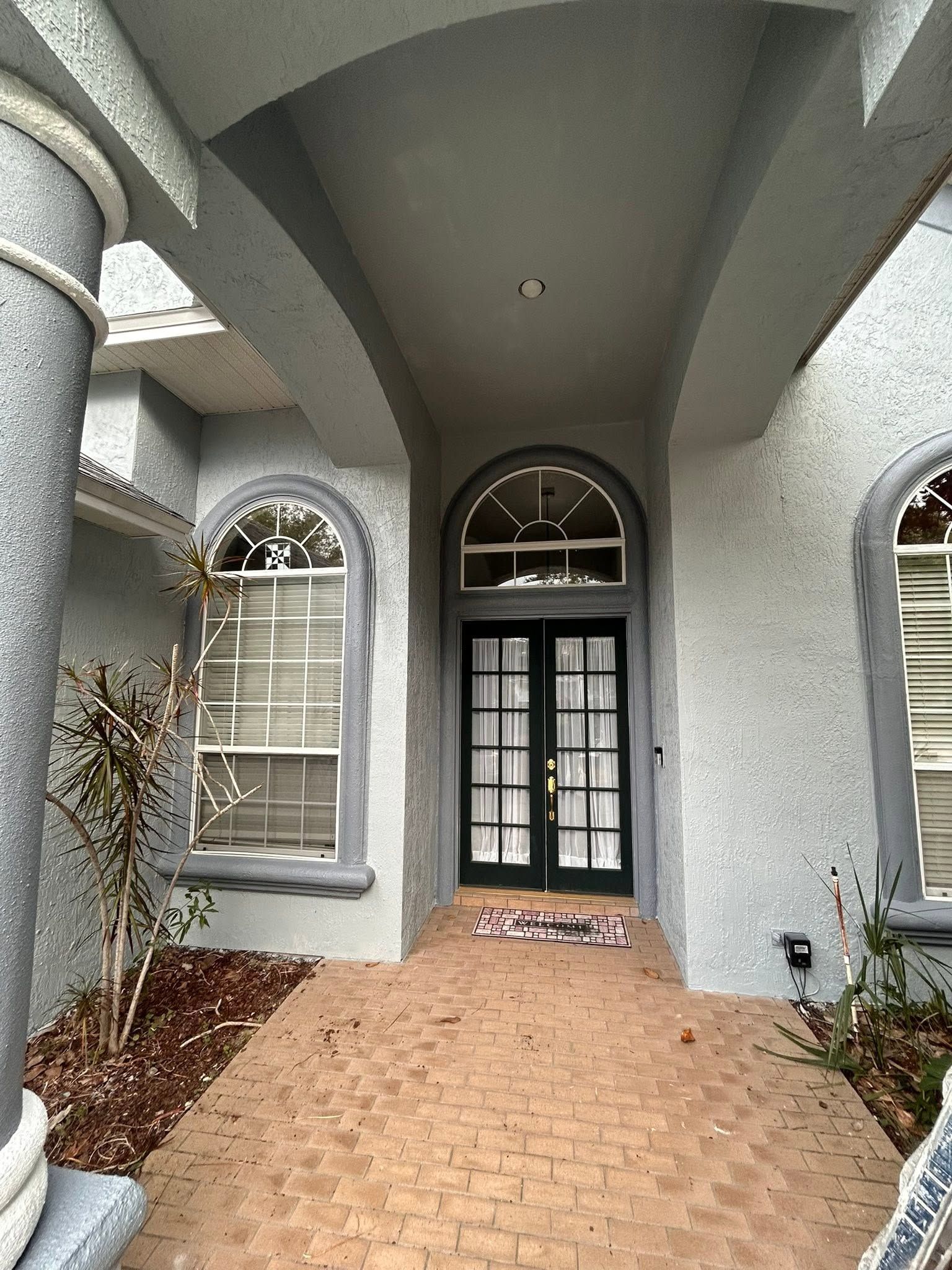 Covered front entryway with arched doorway, gray stucco walls, and brick walkway leading to a glass door