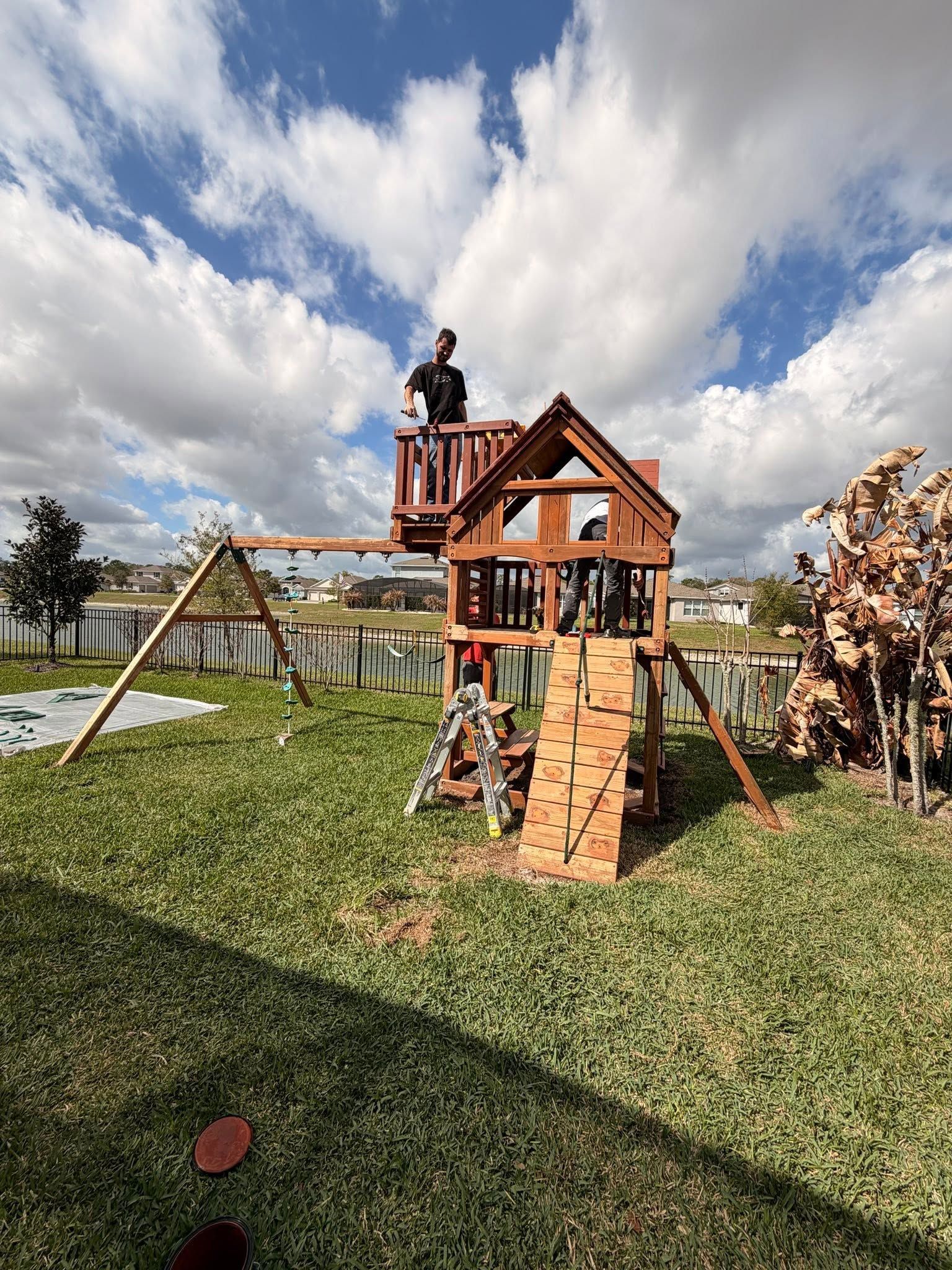 Backyard wooden playhouse with ladder and slide under a cloudy sky, child on the upper platform