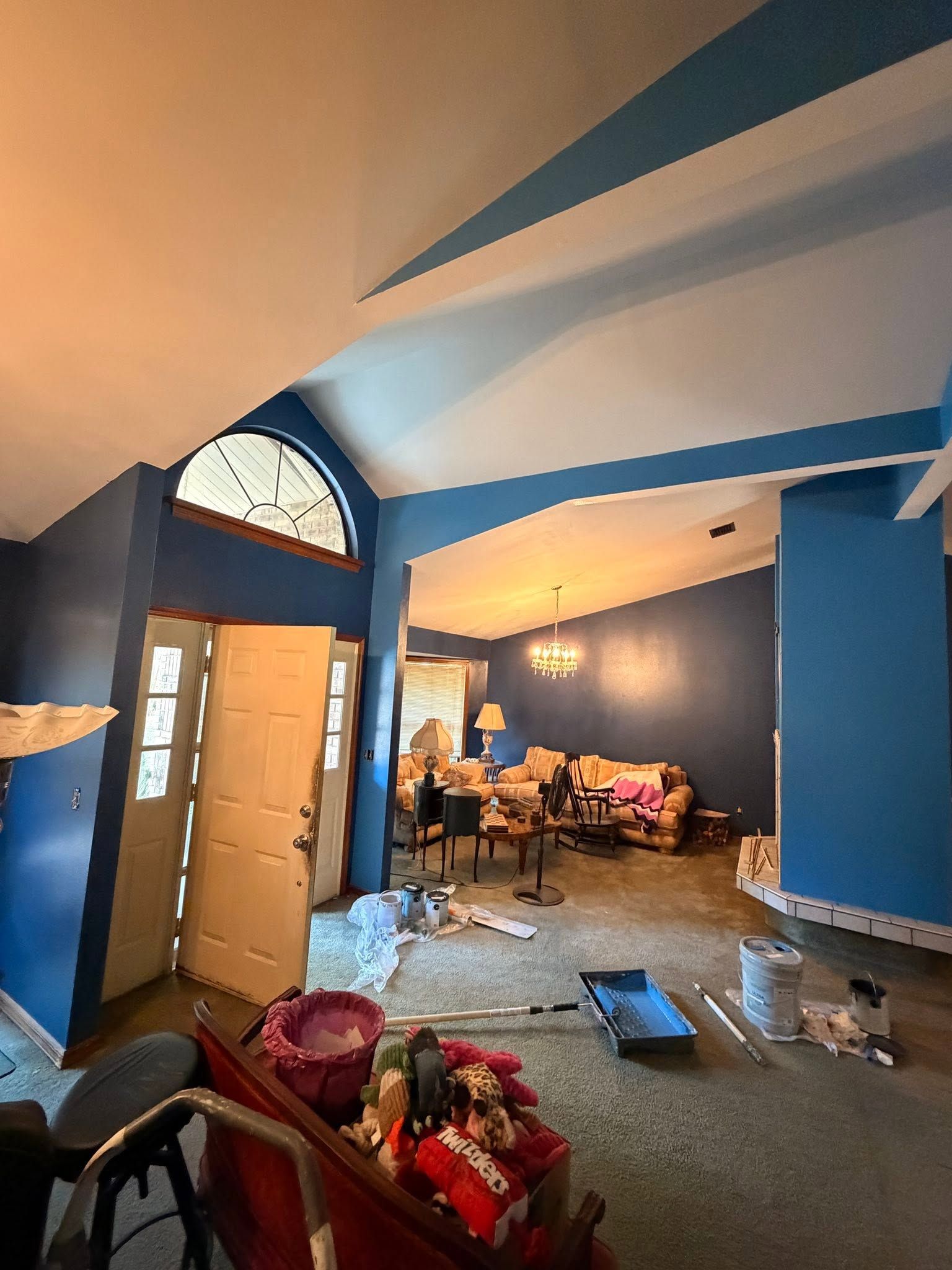 Cluttered living room with exposed beams, open front door, and furniture against blue walls.