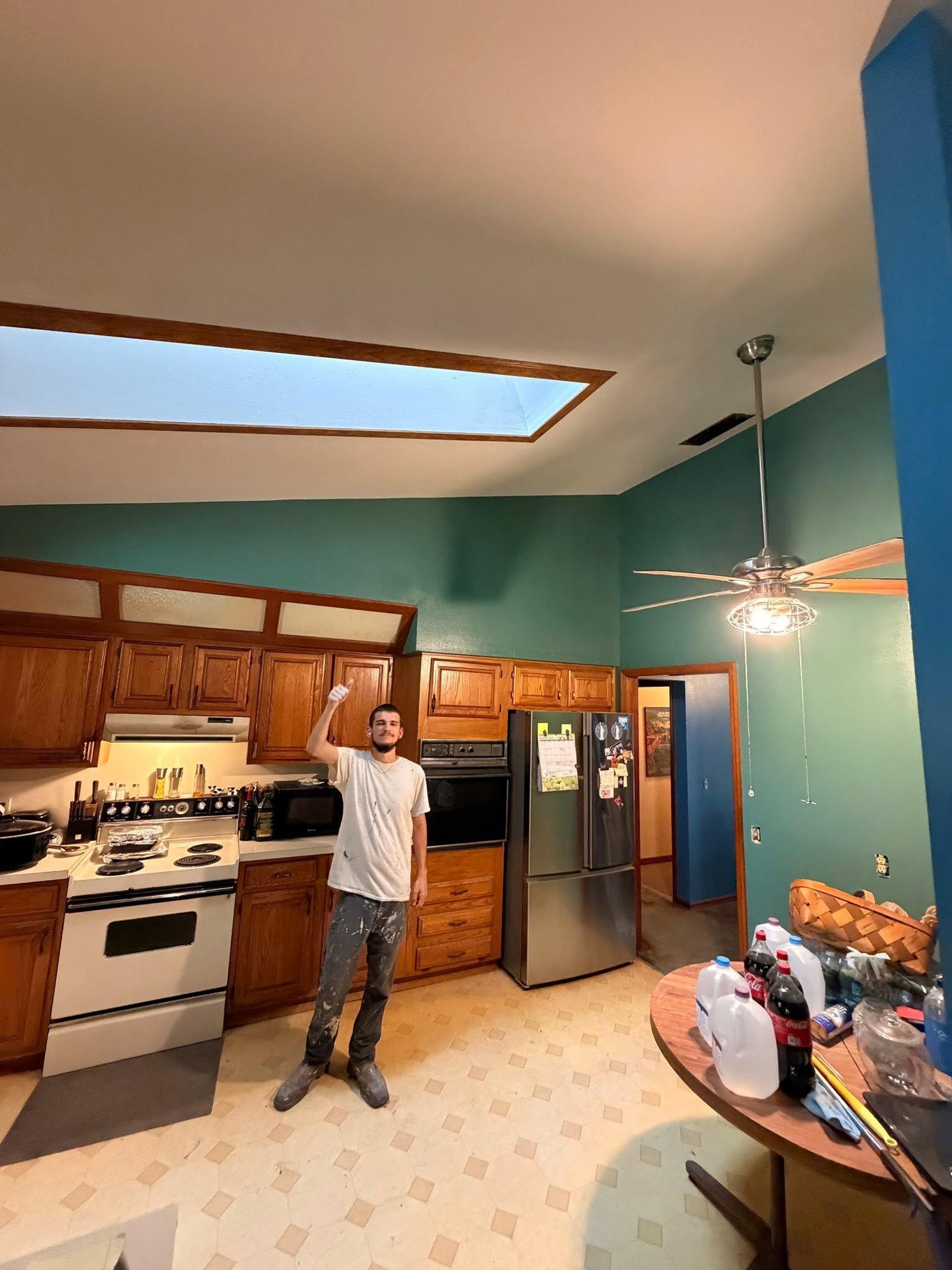 Person standing in a kitchen with wooden cabinets, teal walls, and a stainless-steel fridge