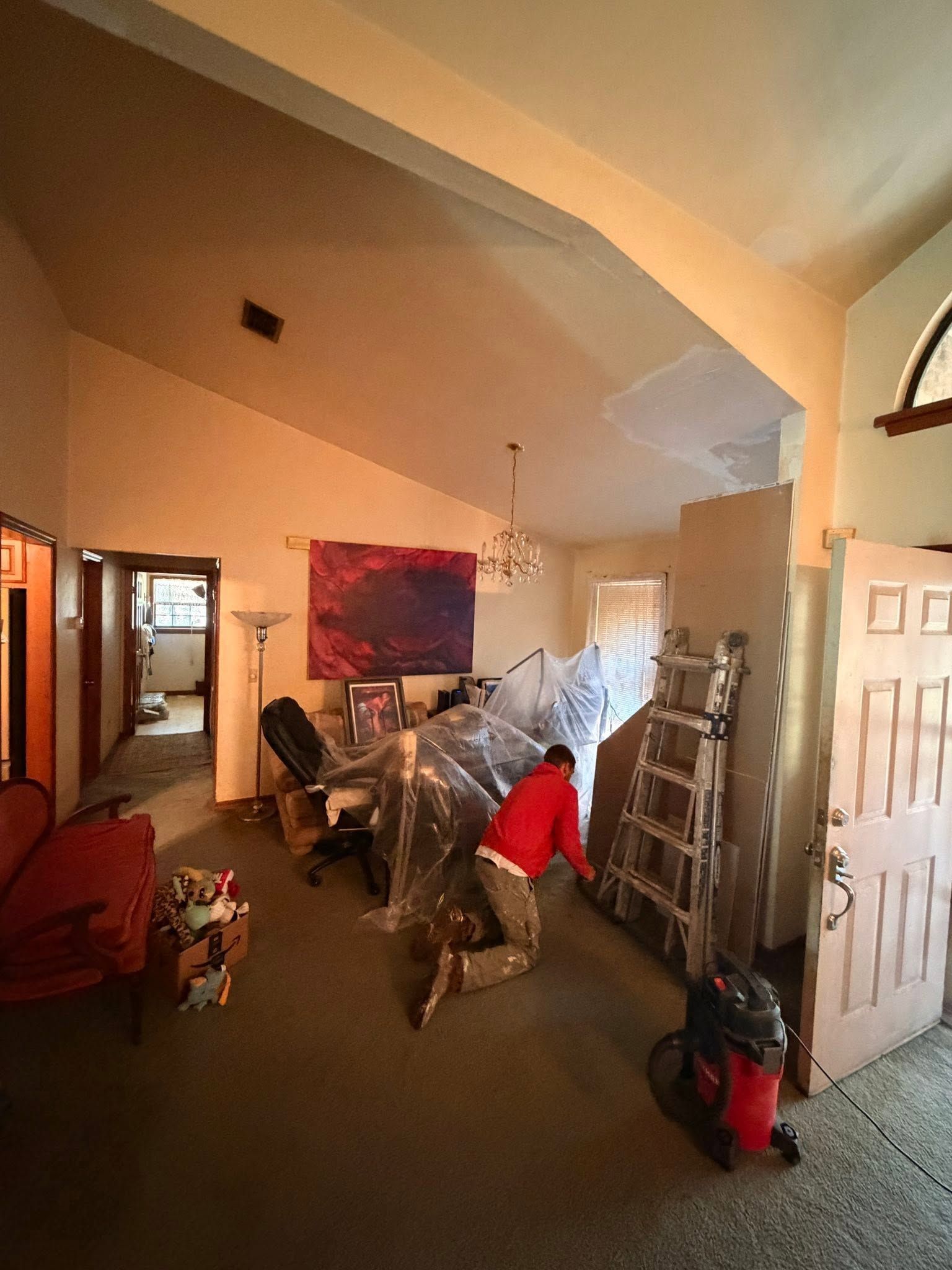 Person in red shirt working in a cluttered room with shelves, chair, and open door.