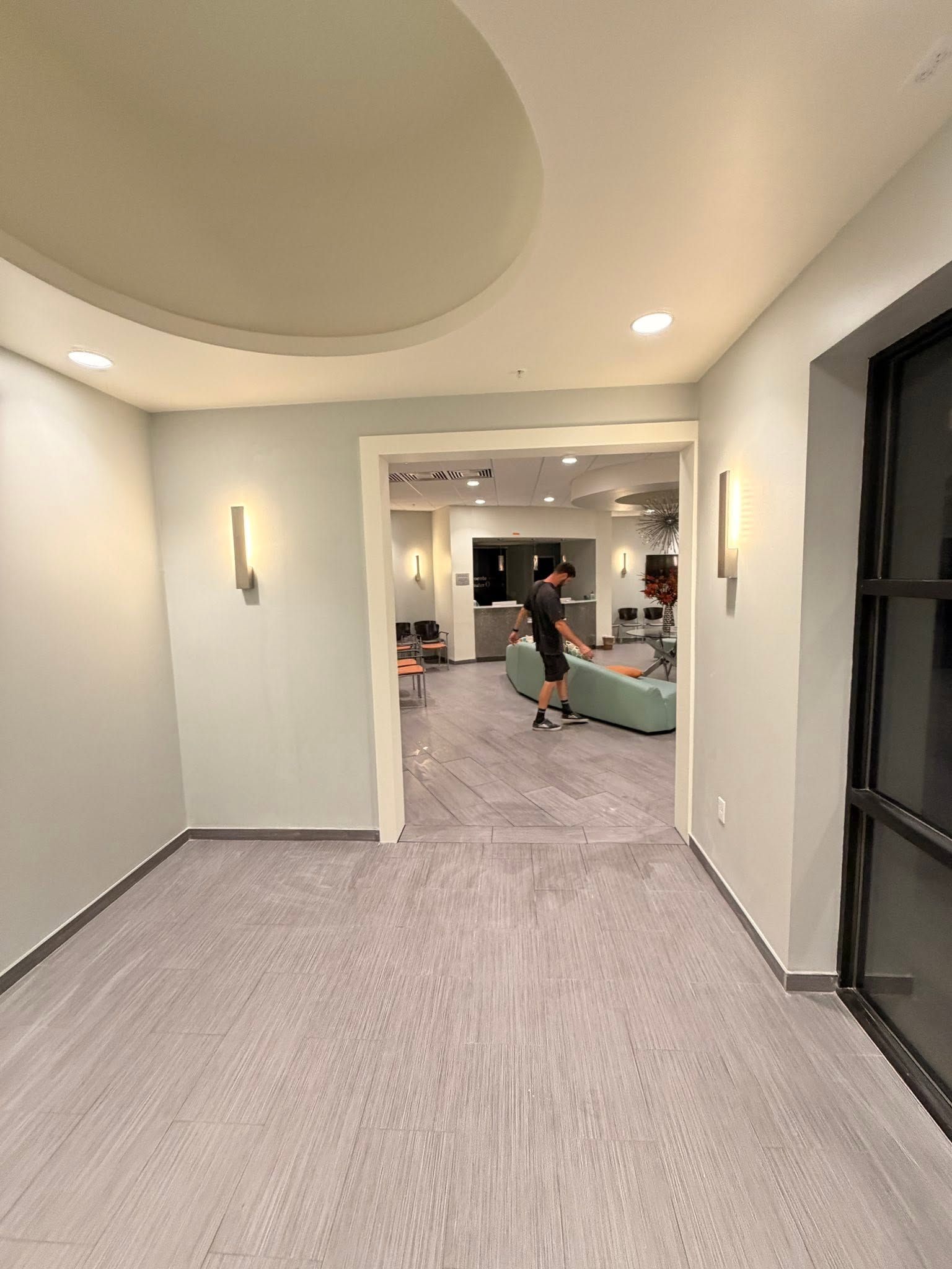 Empty hallway with light carpet and recessed lights leading to a living room with a person and couch.