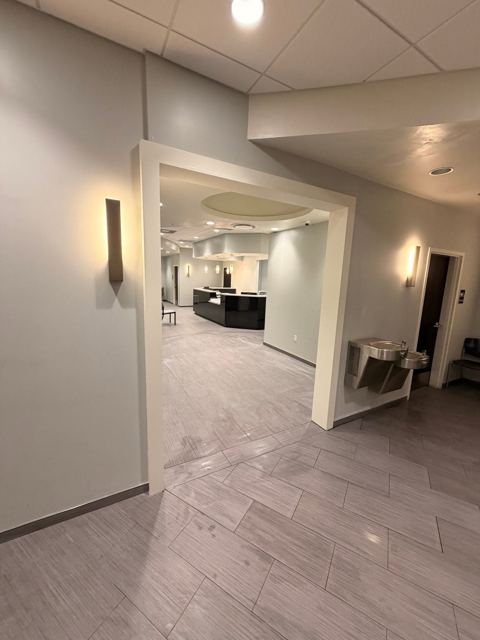 Empty office hallway with beige walls, gray carpet, and wall sconces leading into a bright reception area