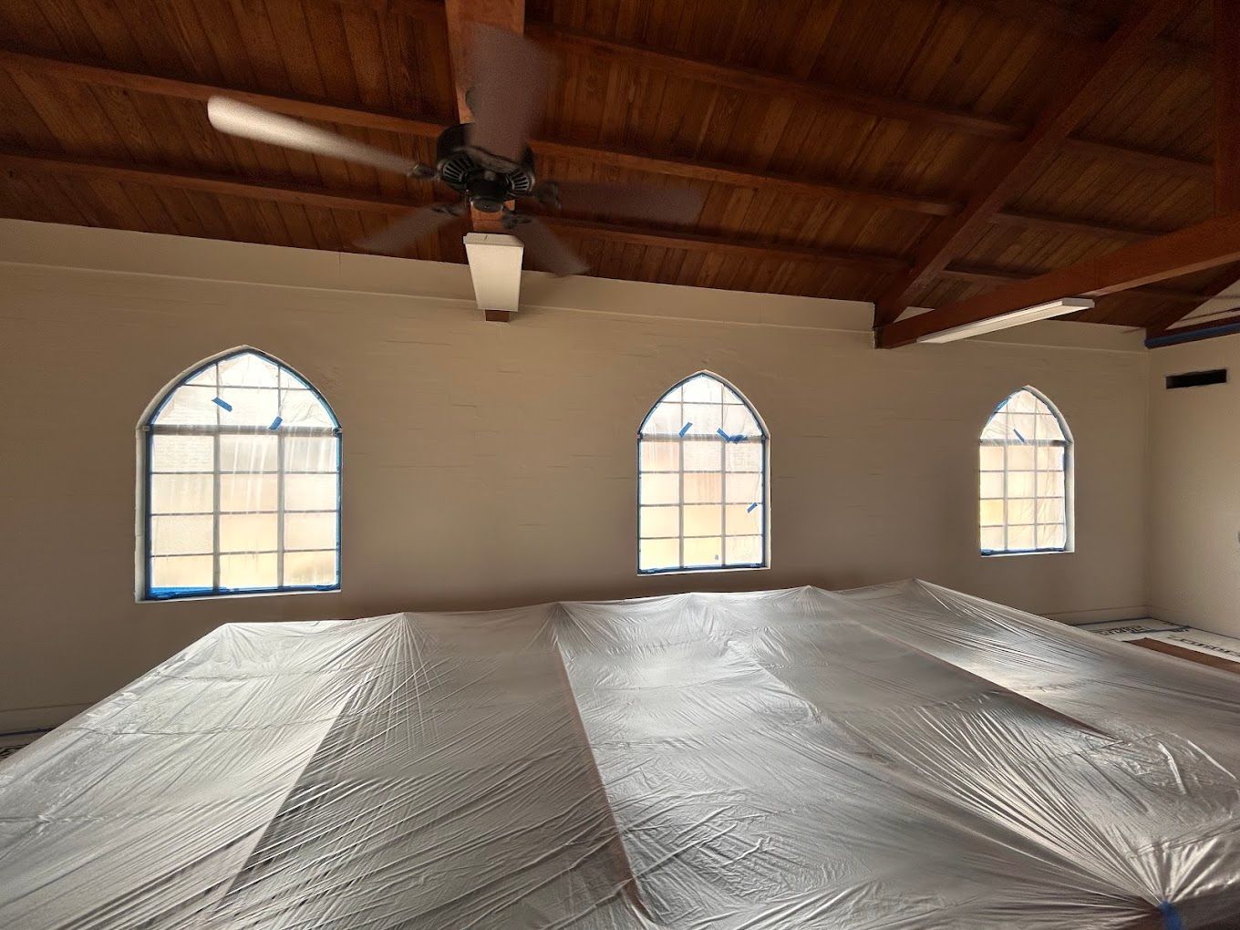 Empty room with wood-beamed ceiling, ceiling fan, arched windows, and plastic-covered floor