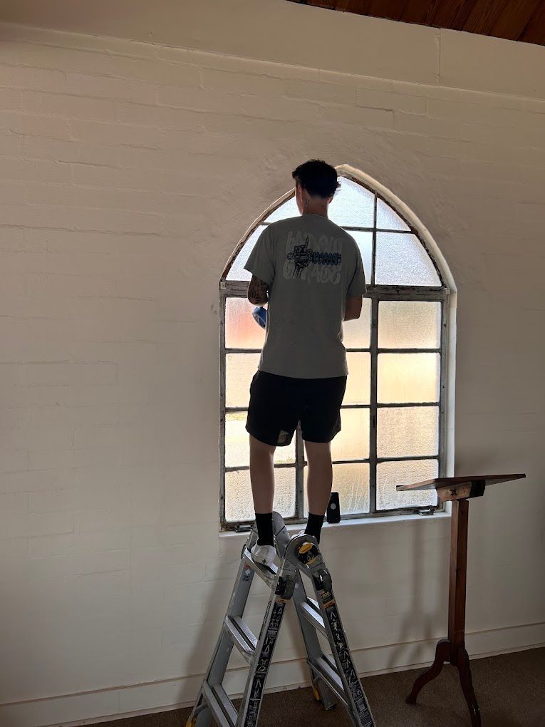 Person standing on a ladder near a large arched window, installing or painting the wall inside a room.