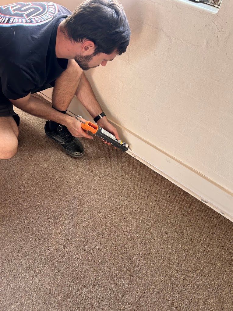 Man applying caulk along baseboard edge in a carpeted room