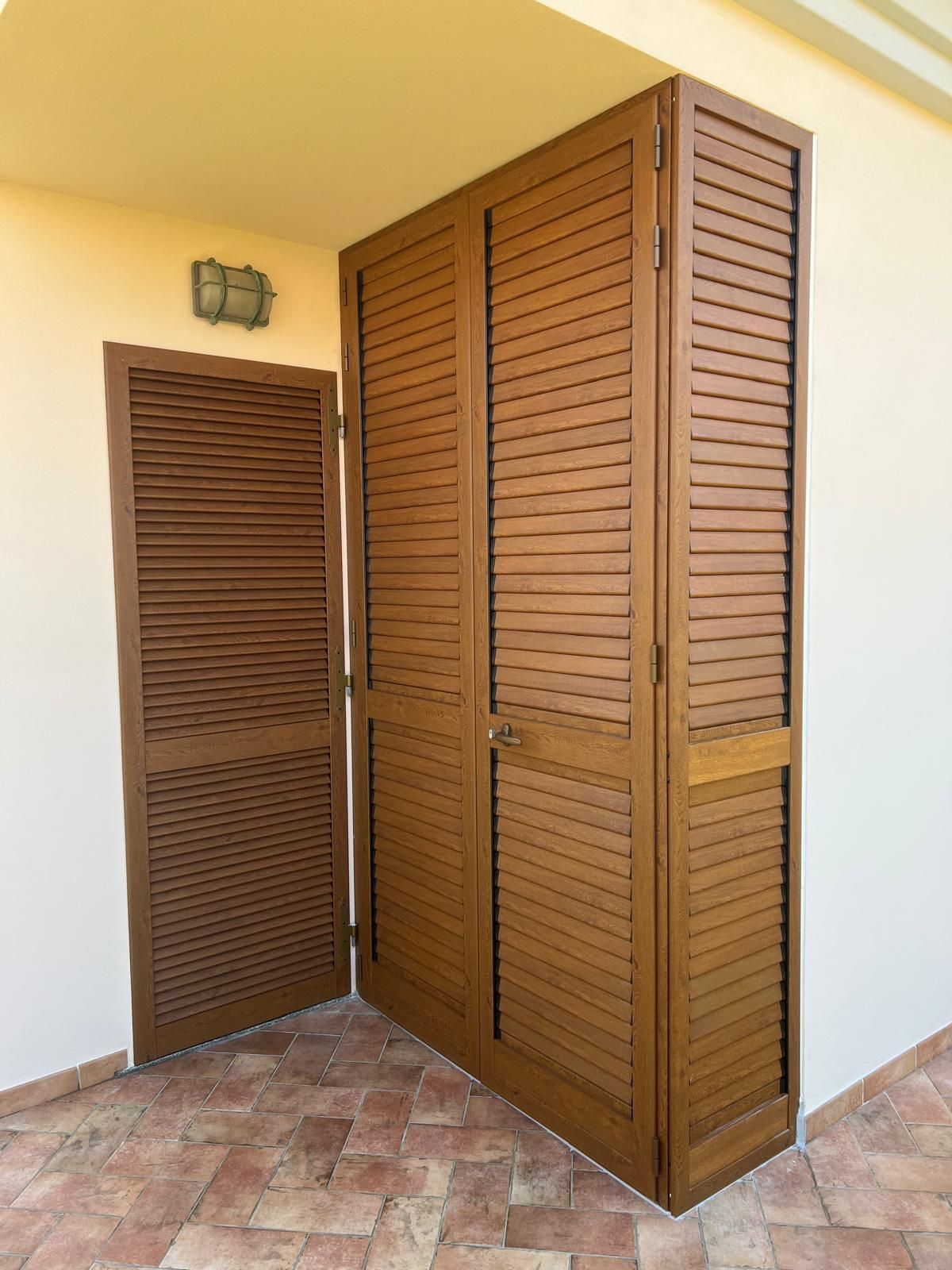 Porte a persiana marroni e una porta singola coordinata, posizionate su un patio rivestito in cotto contro un muro beige.