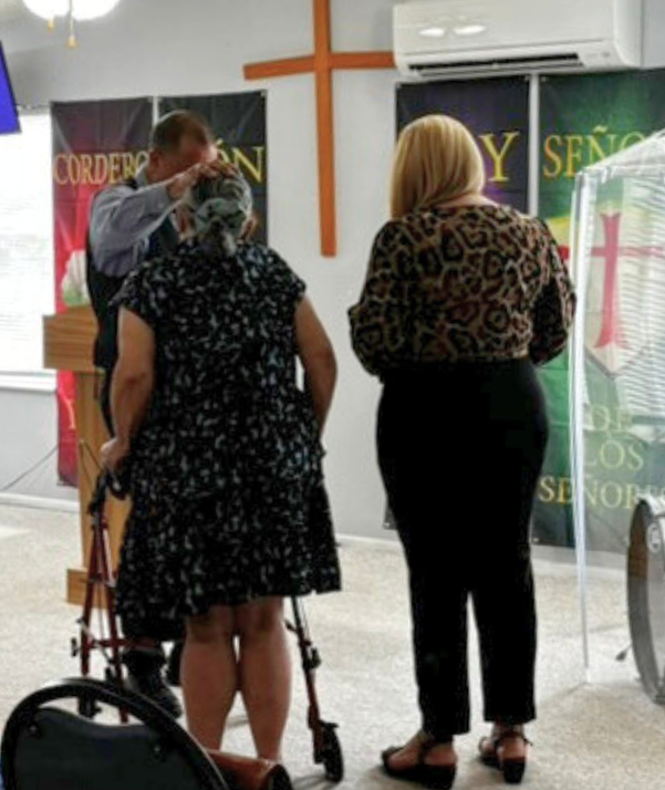 Varias personas de pie en una iglesia cerca de una cruz; se pueden ver personas con andadores y una silla de ruedas.