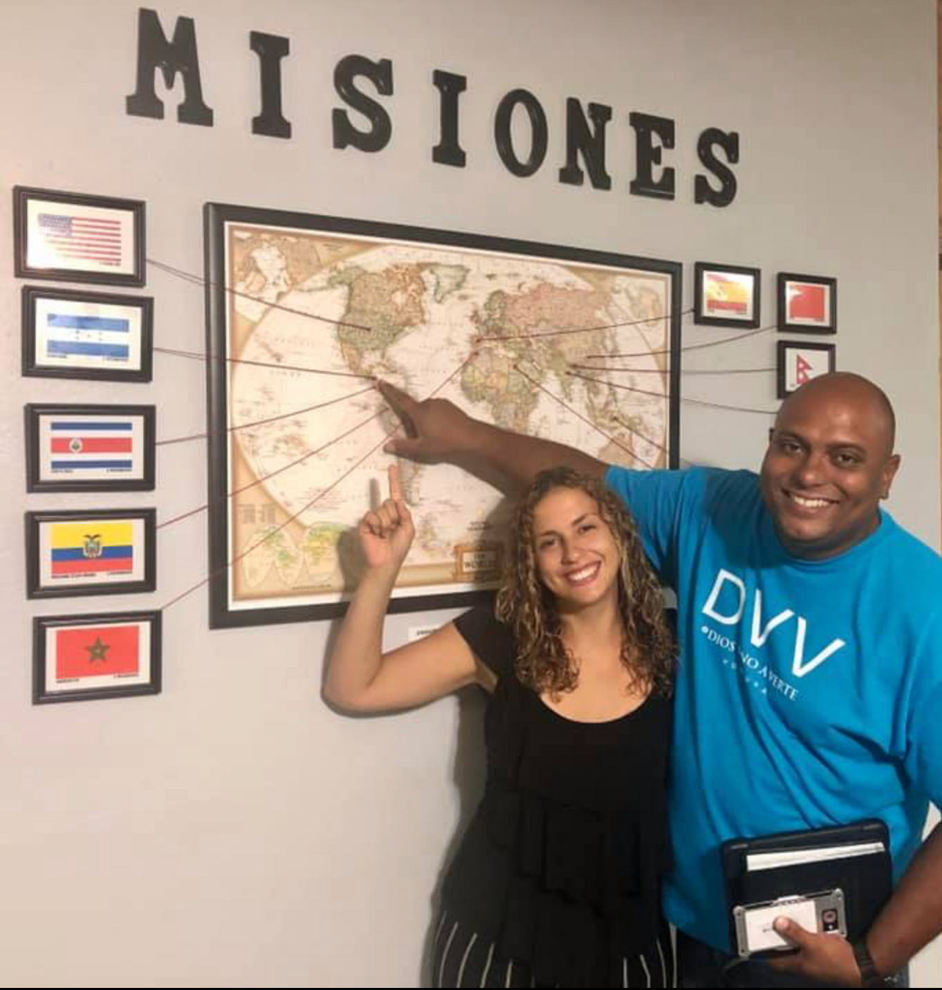 Two smiling people point at a mission map on a wall labeled “MISIONES.”
