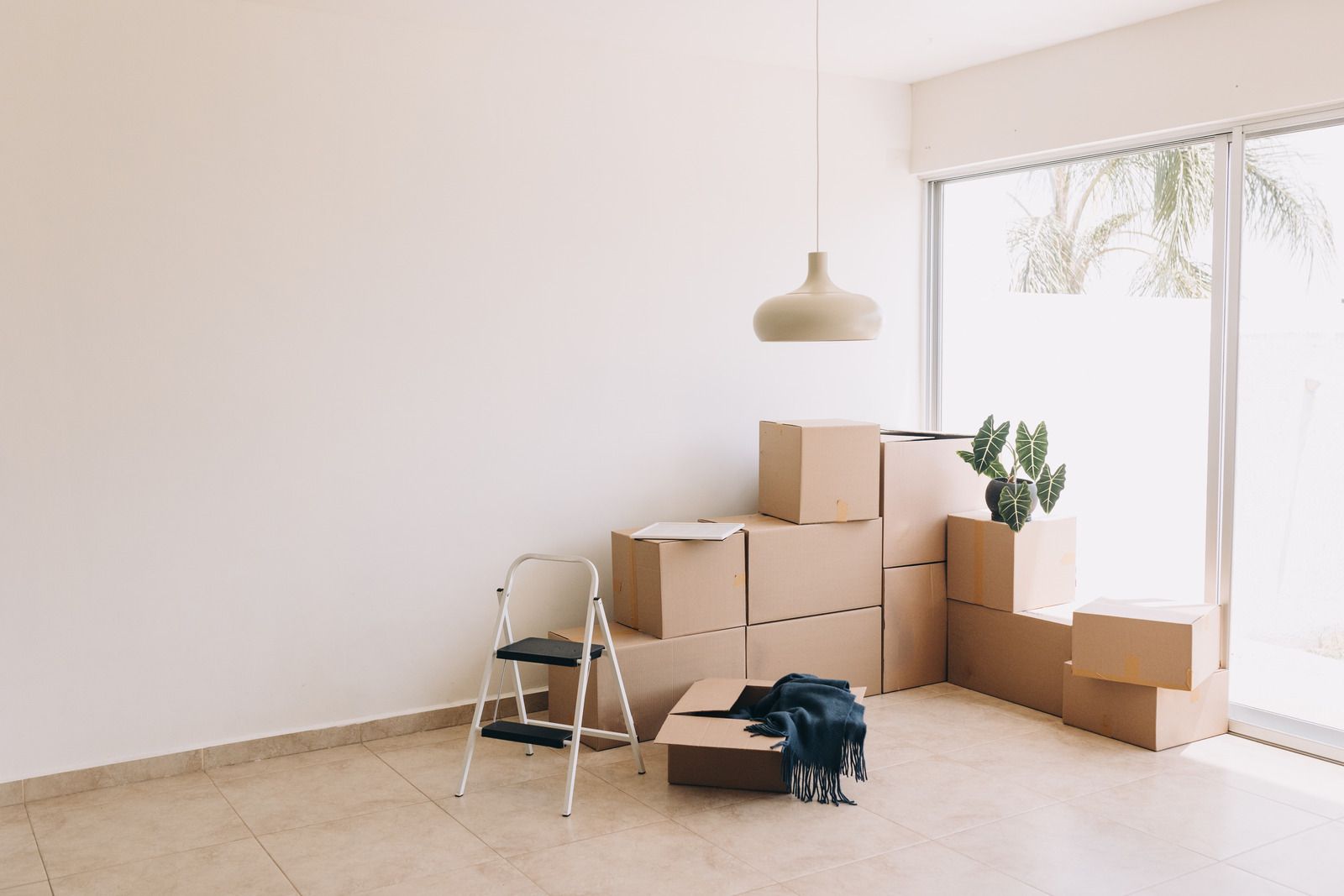 Empty room with stacked moving boxes, a step ladder, and a hanging light fixture.