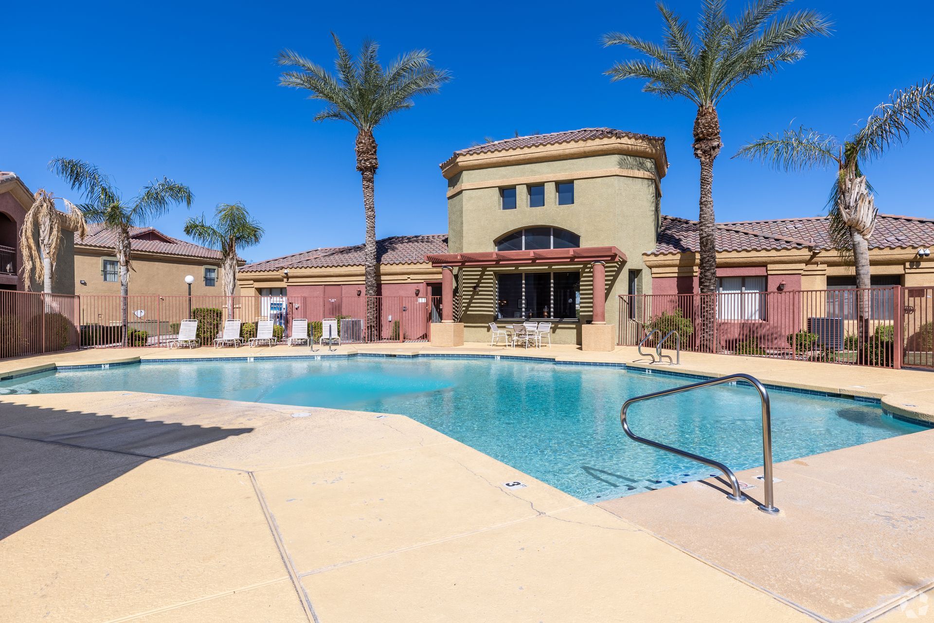 A swimming pool with tan decking, surrounded by palm trees and a Mediterranean-style building under a clear blue sky.