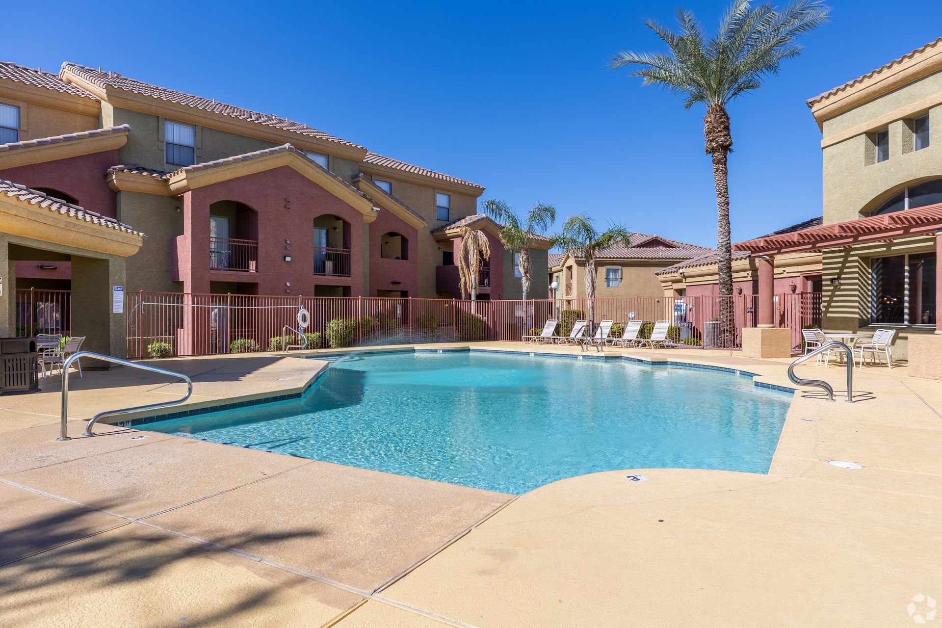 A rectangular swimming pool surrounded by a tan deck with apartment buildings and palm trees under a clear blue sky.