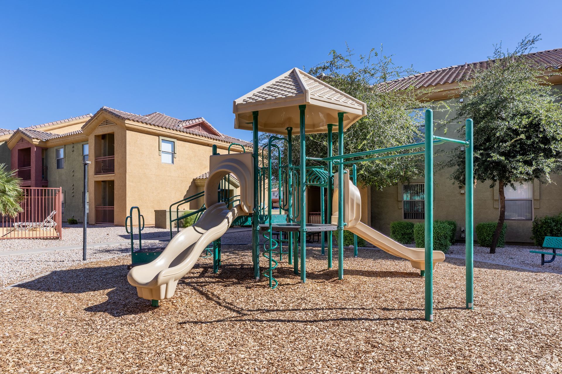 A beige apartment building background with a playground structure featuring a slide, climbing area, and tan wood chips.