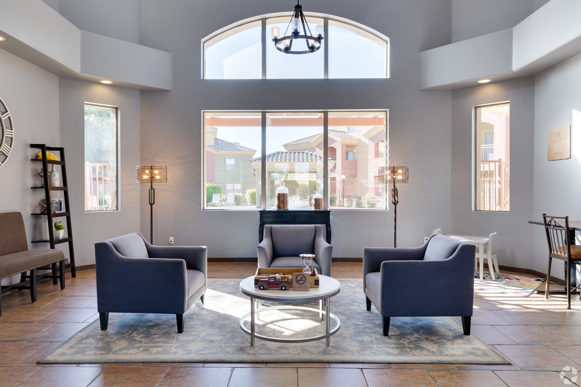 A bright, modern lobby with two blue armchairs, a central coffee table, a tall bookshelf, and large windows.