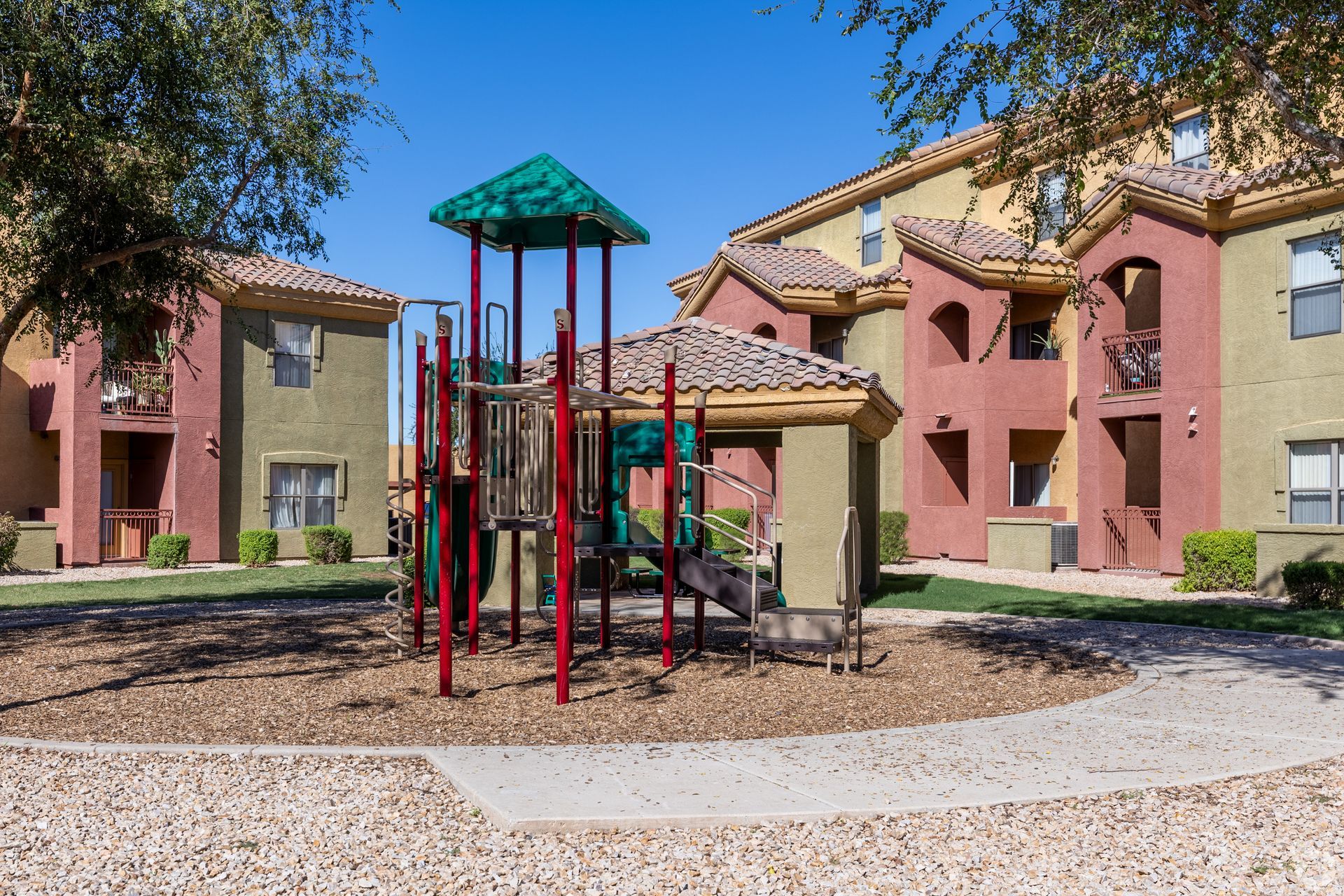 A colorful playground structure stands in a wood-chipped area between two tan and red apartment buildings under a blue sky.