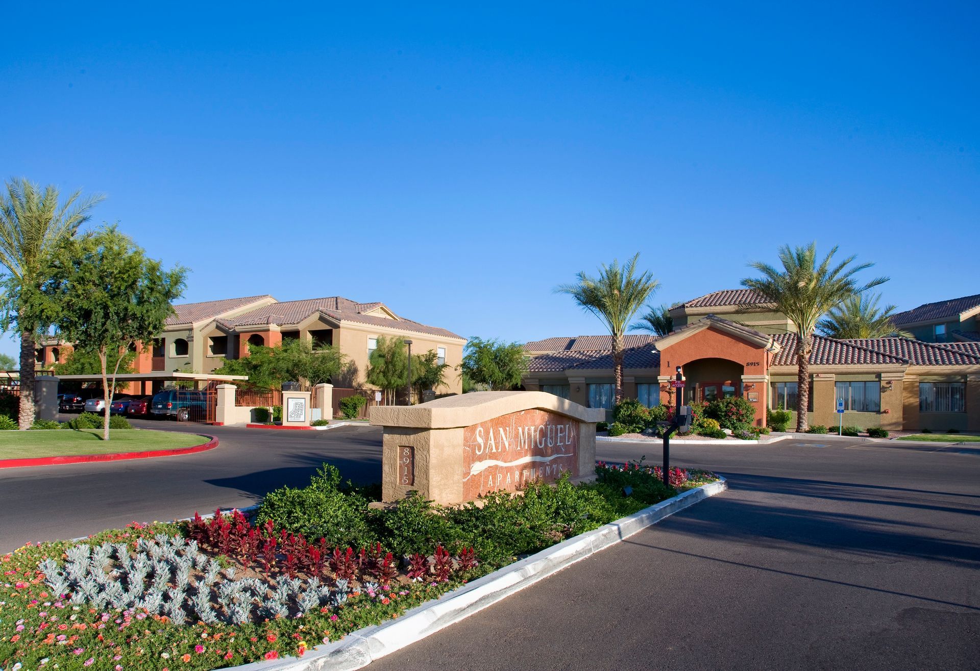 A stone monument sign for San Sonoma at the entrance of an apartment complex with palm trees and buildings under a blue sky.