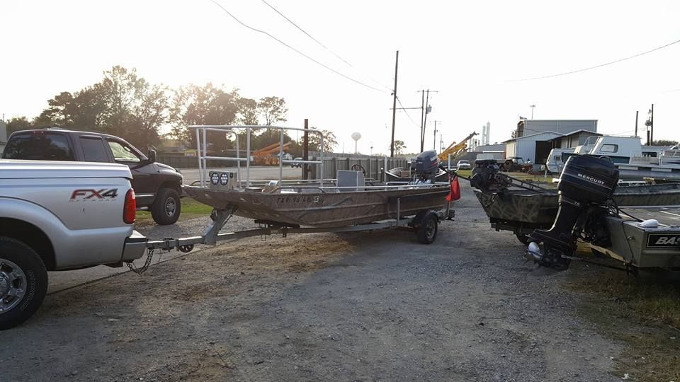 Truck towing a camouflage boat on a trailer, parked on gravel next to another boat. Outdoors setting.