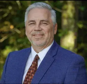 Portrait of a smiling man in a blue suit and patterned tie against a blurred outdoor background