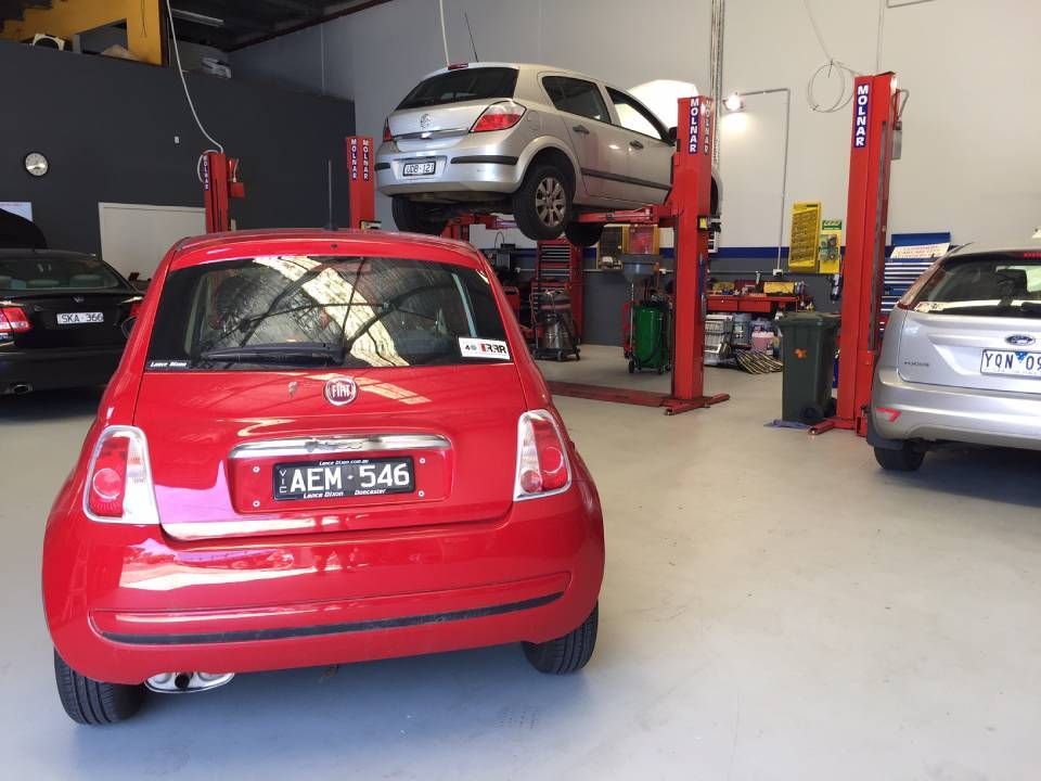 Red hatchback in an auto repair shop with a silver car lifted on a hoist in the background