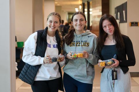 Three individuals stand in a hallway, smiling and holding small, clear containers of food.