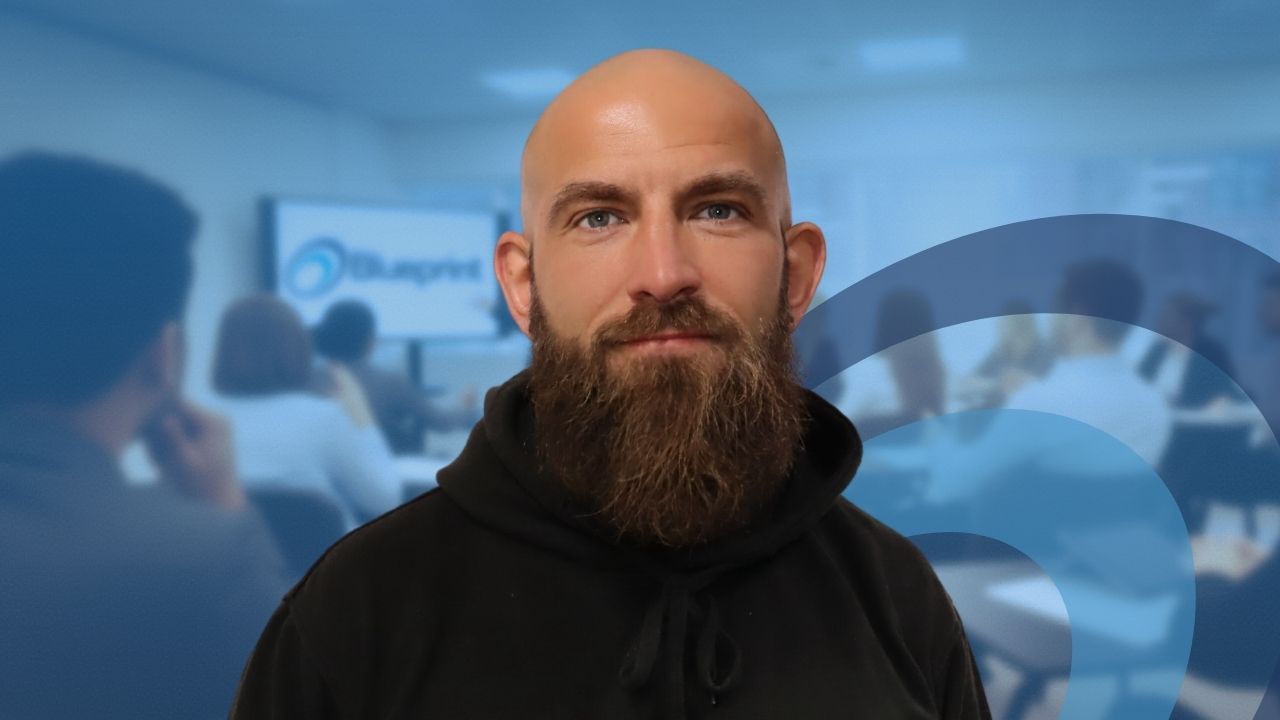 Bald man with a large beard, black hoodie, and blue background with a blurred classroom setting.