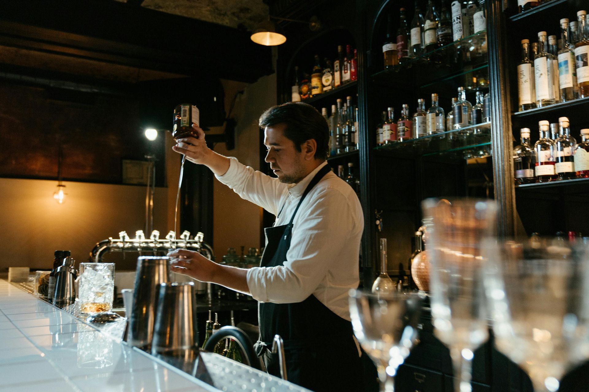 Bartender pouring drink into shaker behind a bar with liquor bottles on shelves.