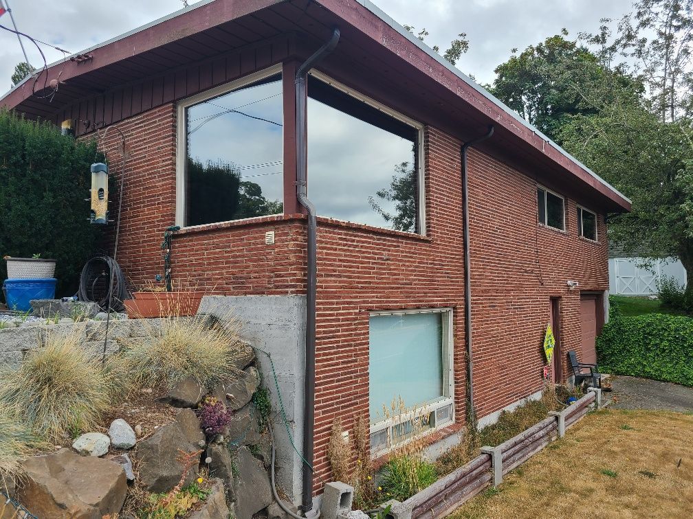 Brick house with large windows and sloping yard. Brown siding, a concrete foundation, and a green yard are visible.