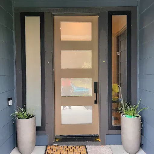 Front door with sidelights, tan, four square panes, gray frame, and two tall planters with greenery.