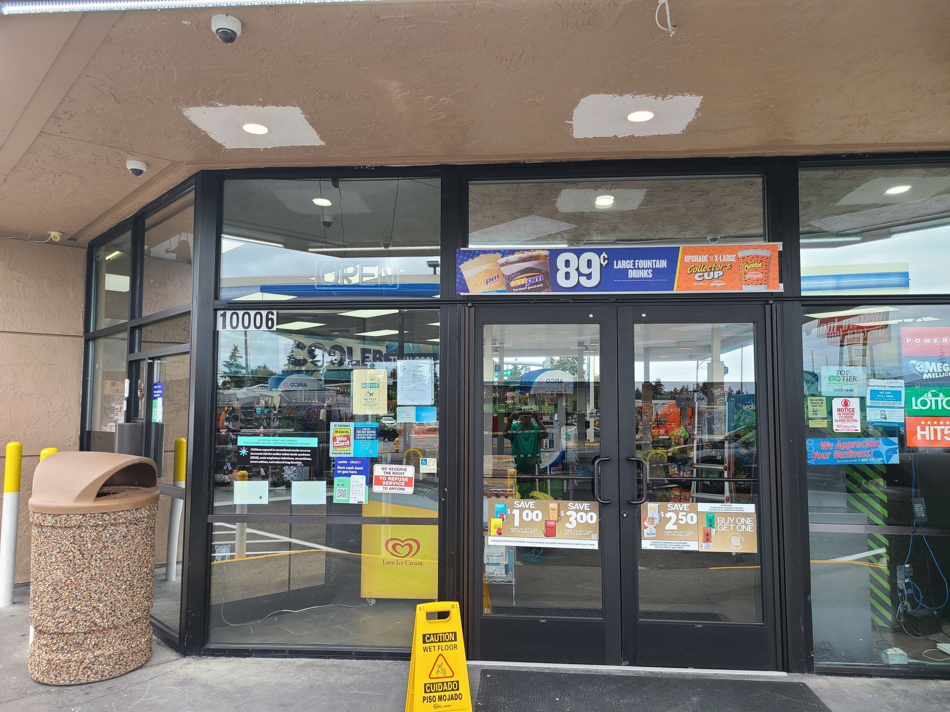 Exterior view of a convenience store with glass doors, various signs, and a trash can on the left.