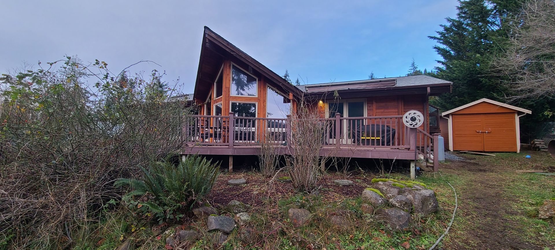 Wooden cabin with a-frame roof and deck. Small shed to the side, set in a natural landscape.