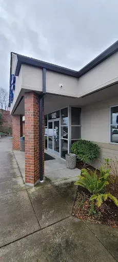 Exterior of a building with glass doors, brick pillars, and landscaping on a cloudy day.
