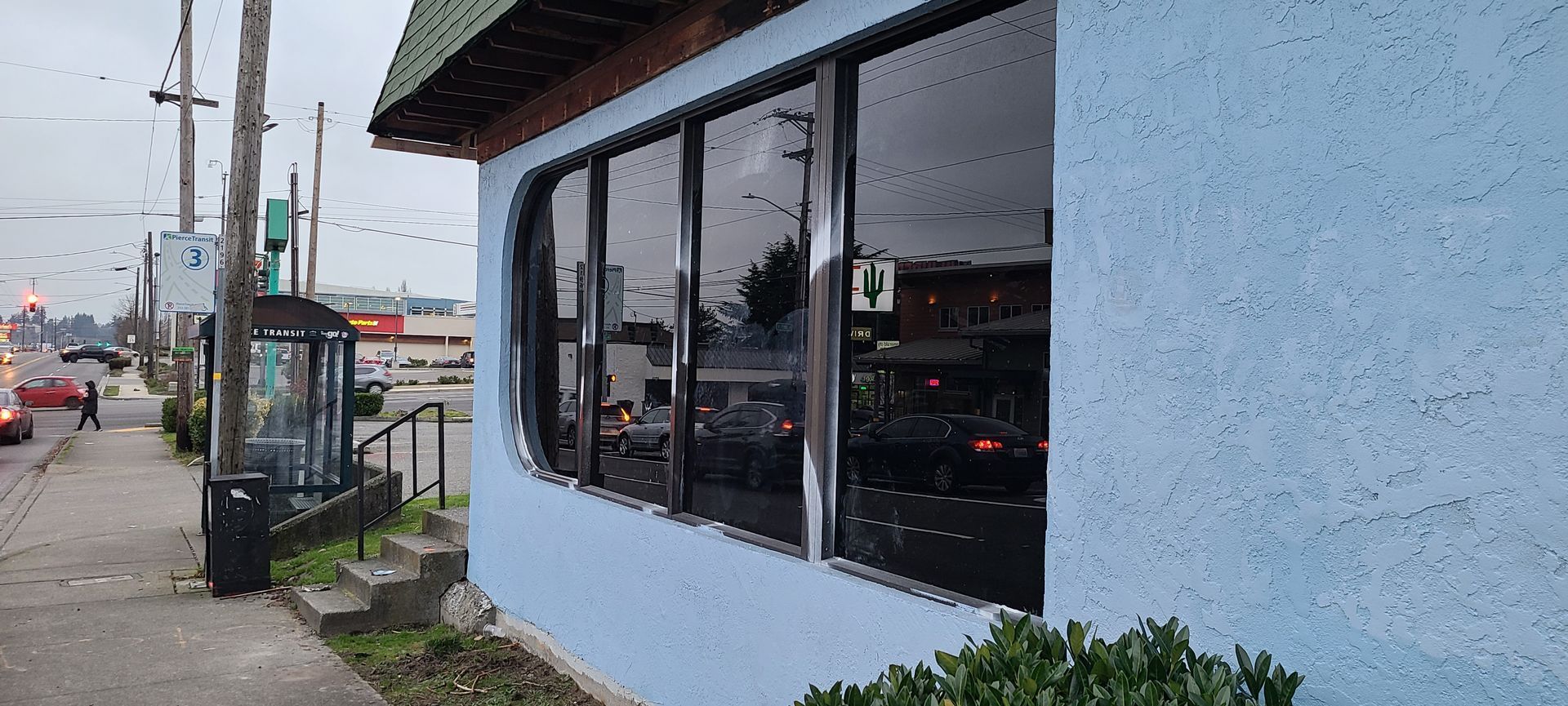Blue building with large windows, bus stop on the sidewalk, and a street in the background.