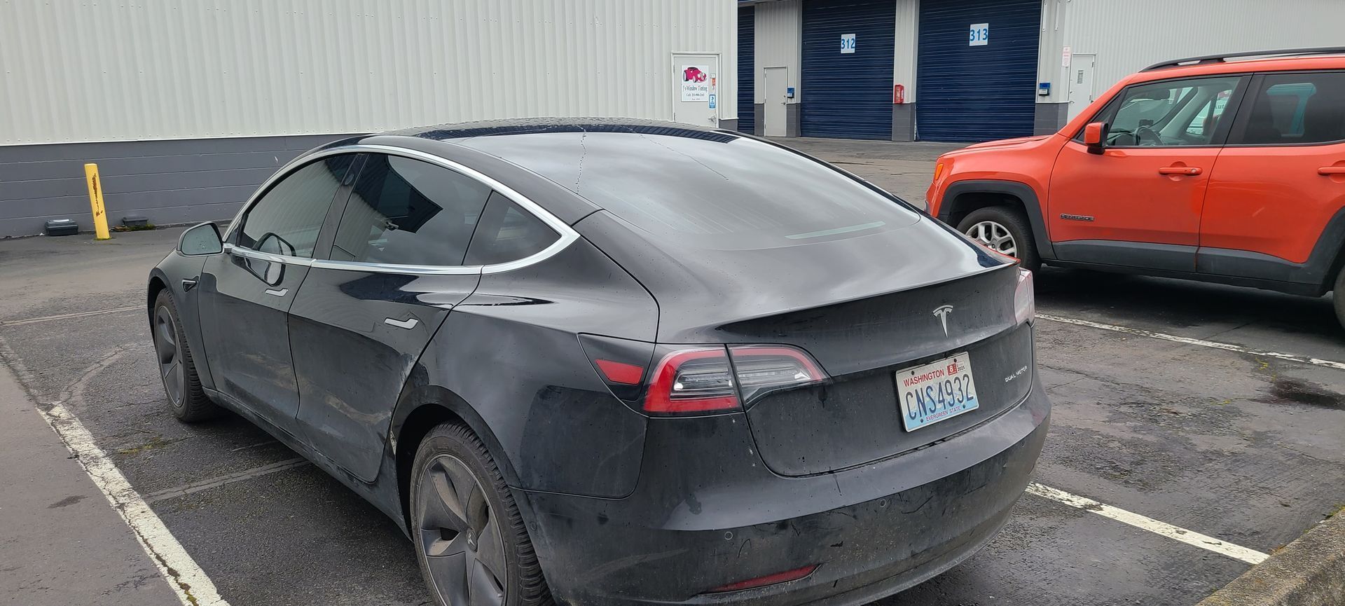 Black Tesla Model 3 parked in a parking lot with an orange SUV next to it.