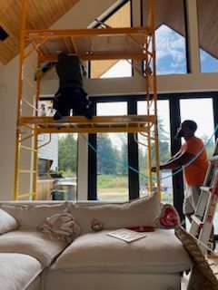 Two workers installing something near high windows, one on a scaffold, other on a ladder. Cat on couch below.