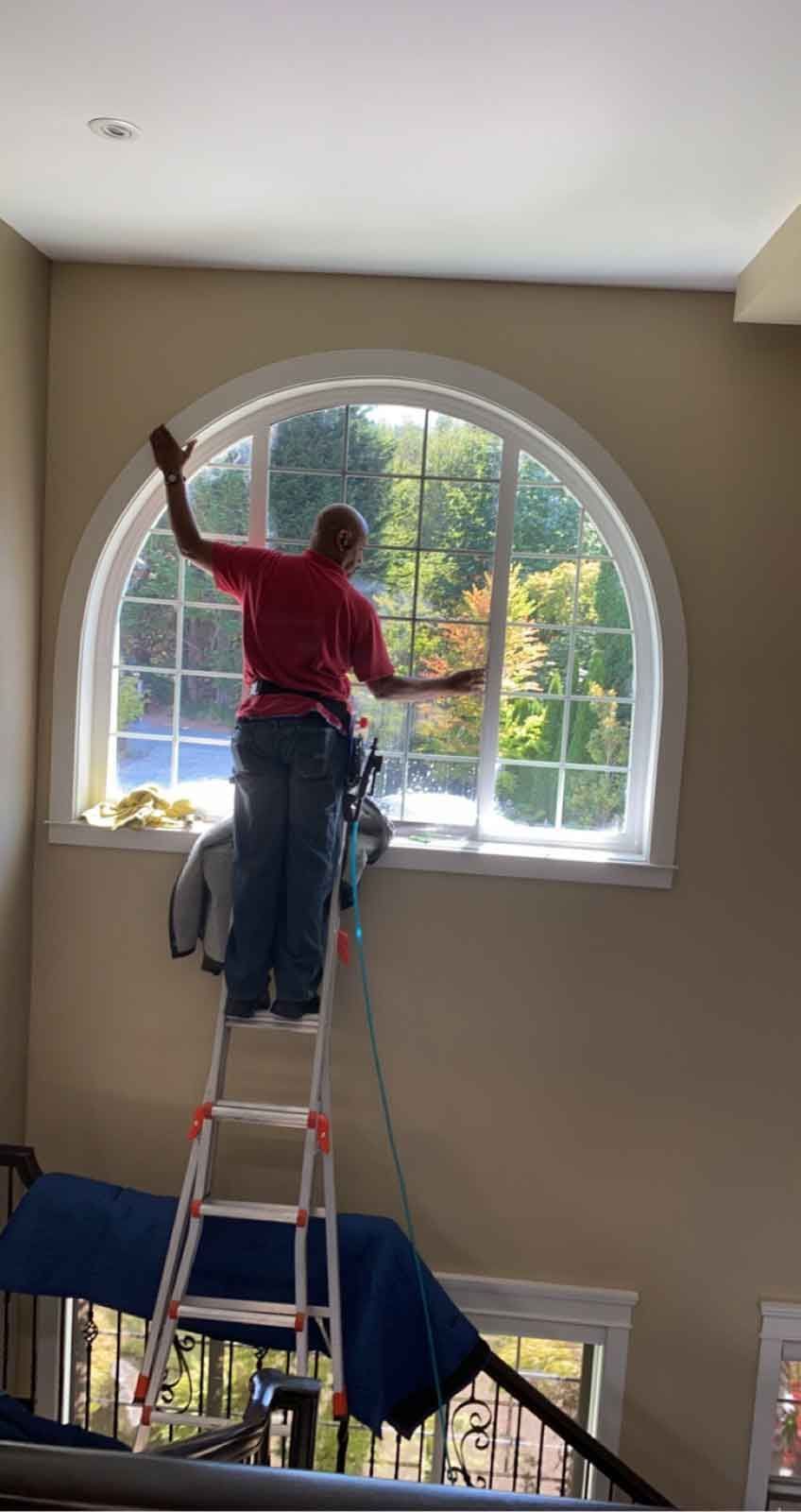 Man cleaning a large arched window from a ladder. He is in a home, reaching up to the window.
